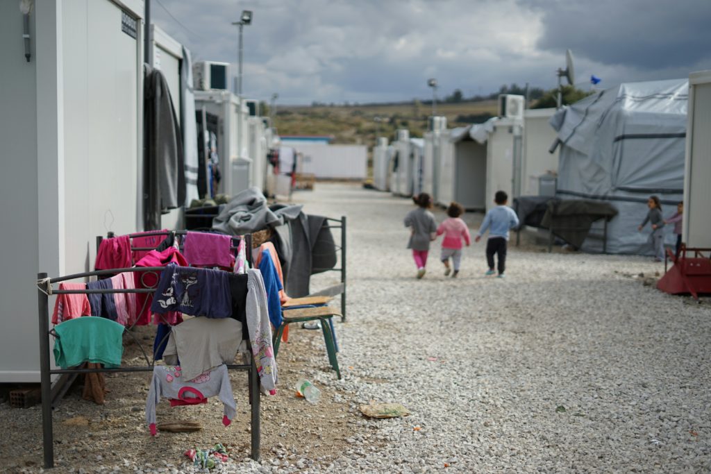 Viser konturene av en flyktningleir, tre små barn går med ryggen til og holder hverandre i hånden. 